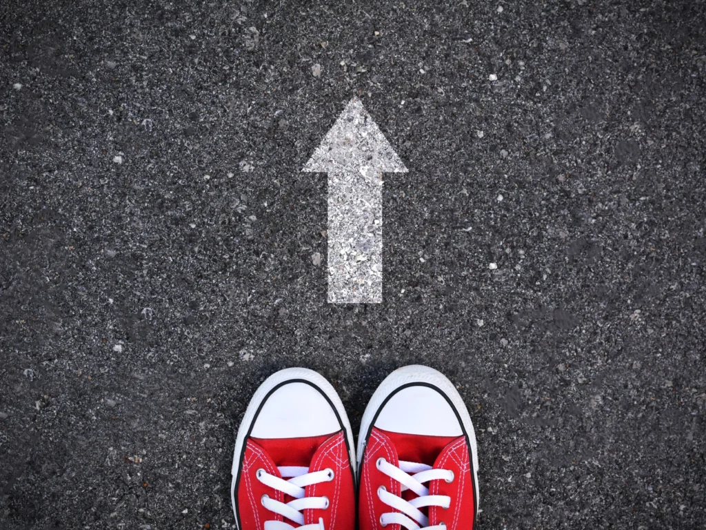 A pair of red Converse sneakers stand on a bitchumen road. A white arrow painted on the road points away from the shoes.