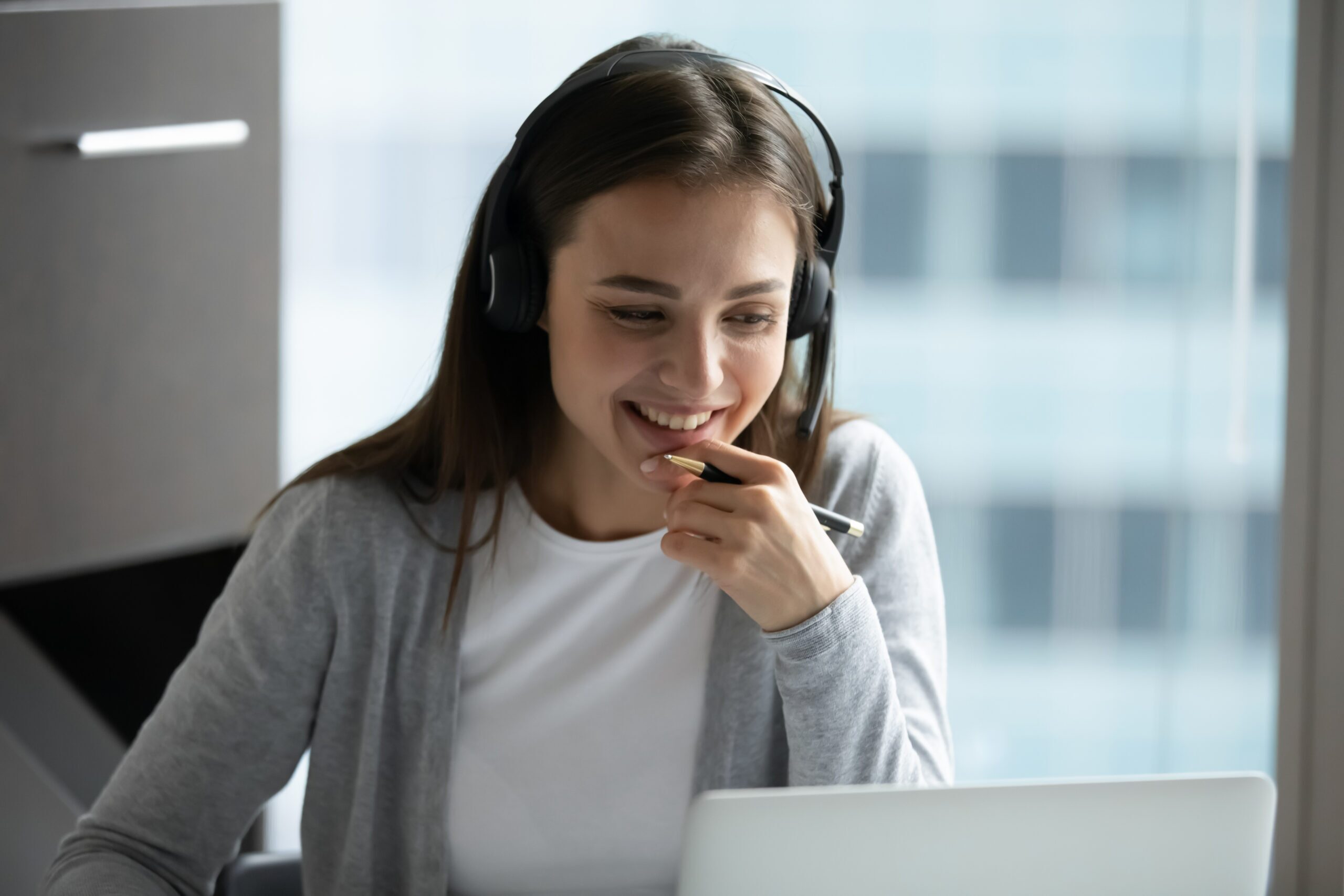 Woman wearing a headset seated in an office looks at laptop while smiling and holding a pen