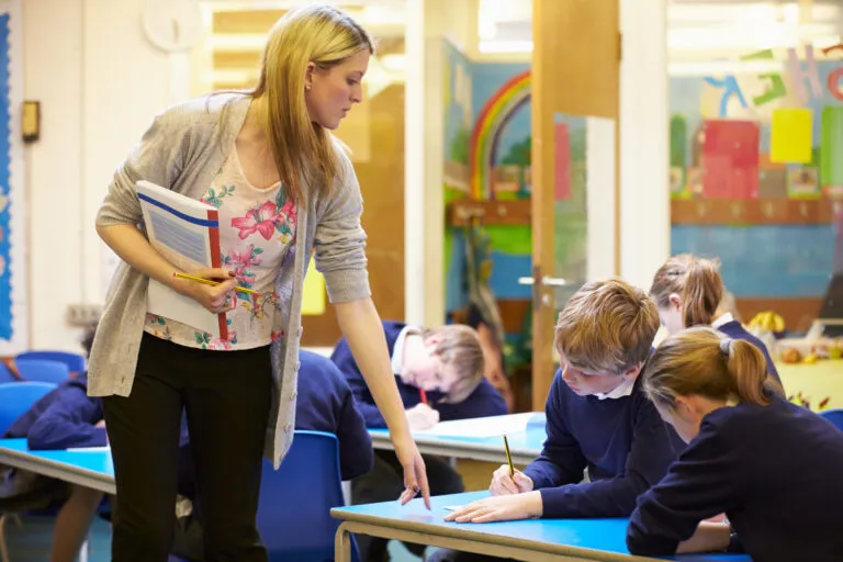 In a classroom, a teacher supervises a class of primary school students who are writing