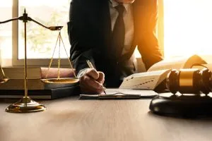 A lawyer in a suit signs a document while consulting a large reference book. On the desk are scales and a gavel.