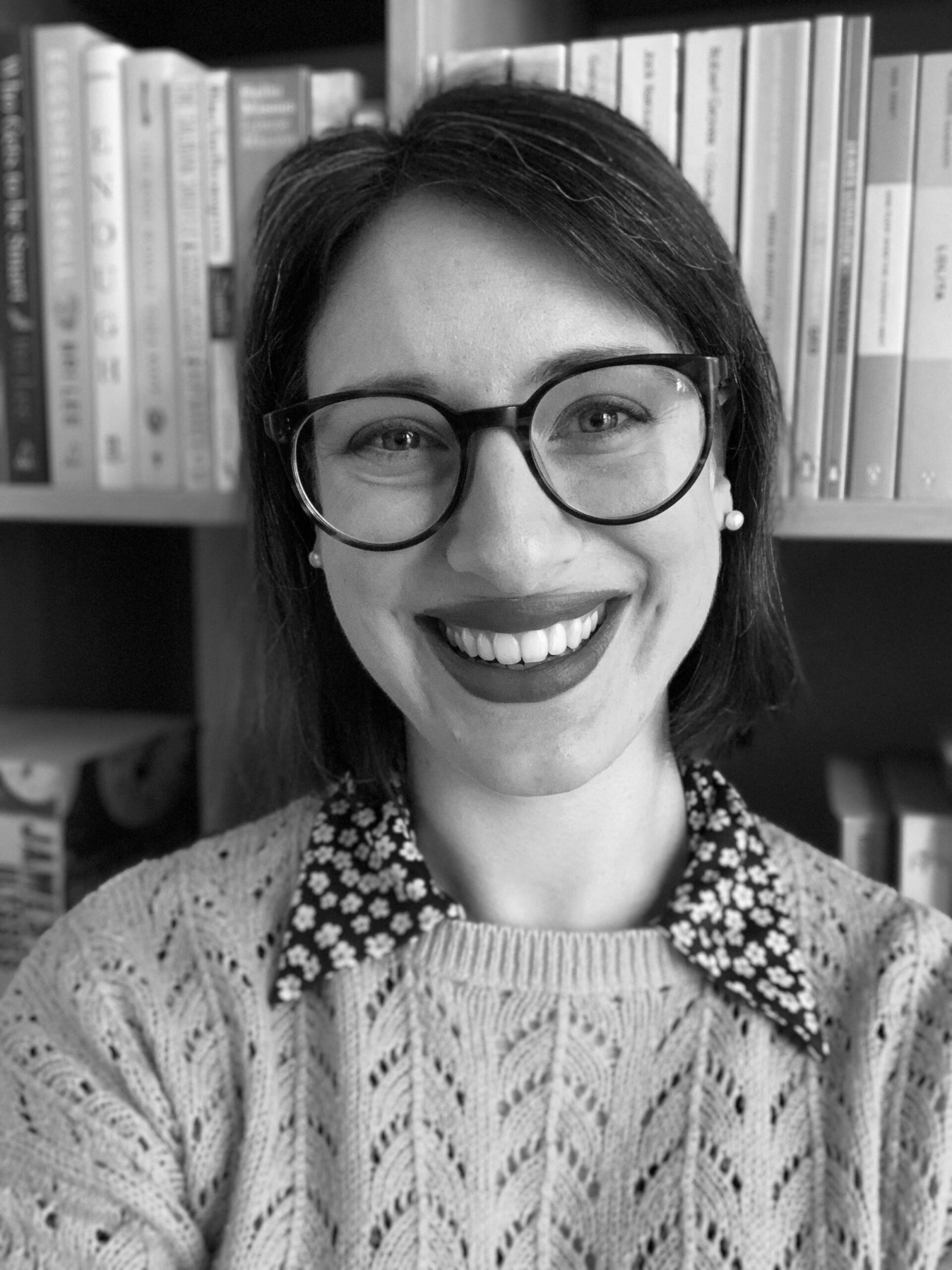 Accredited editor Emily Halloran smiles at a camera. She is wearing large reading glasses, a sweater and a collared shirt.