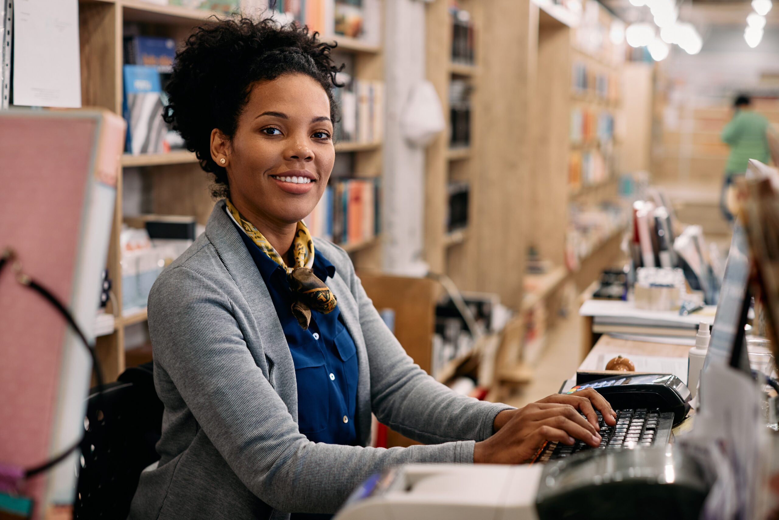 A librarian sits at her desk, working on her computer in a busy local government library