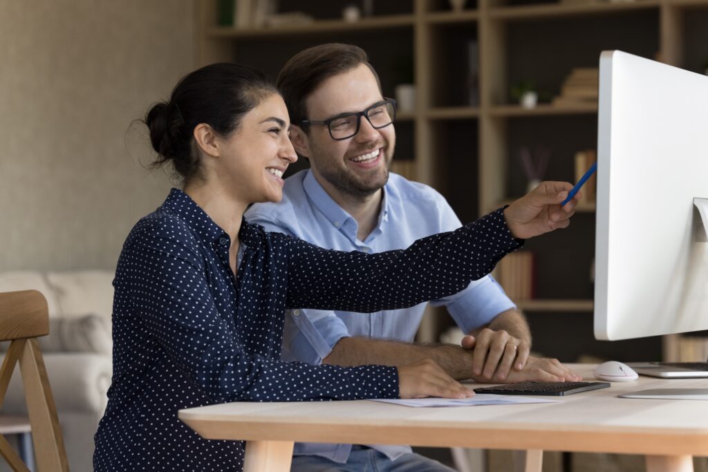 Two work colleagues sit at a desk looking at a computer screen together, while one person points to the screen using her pen