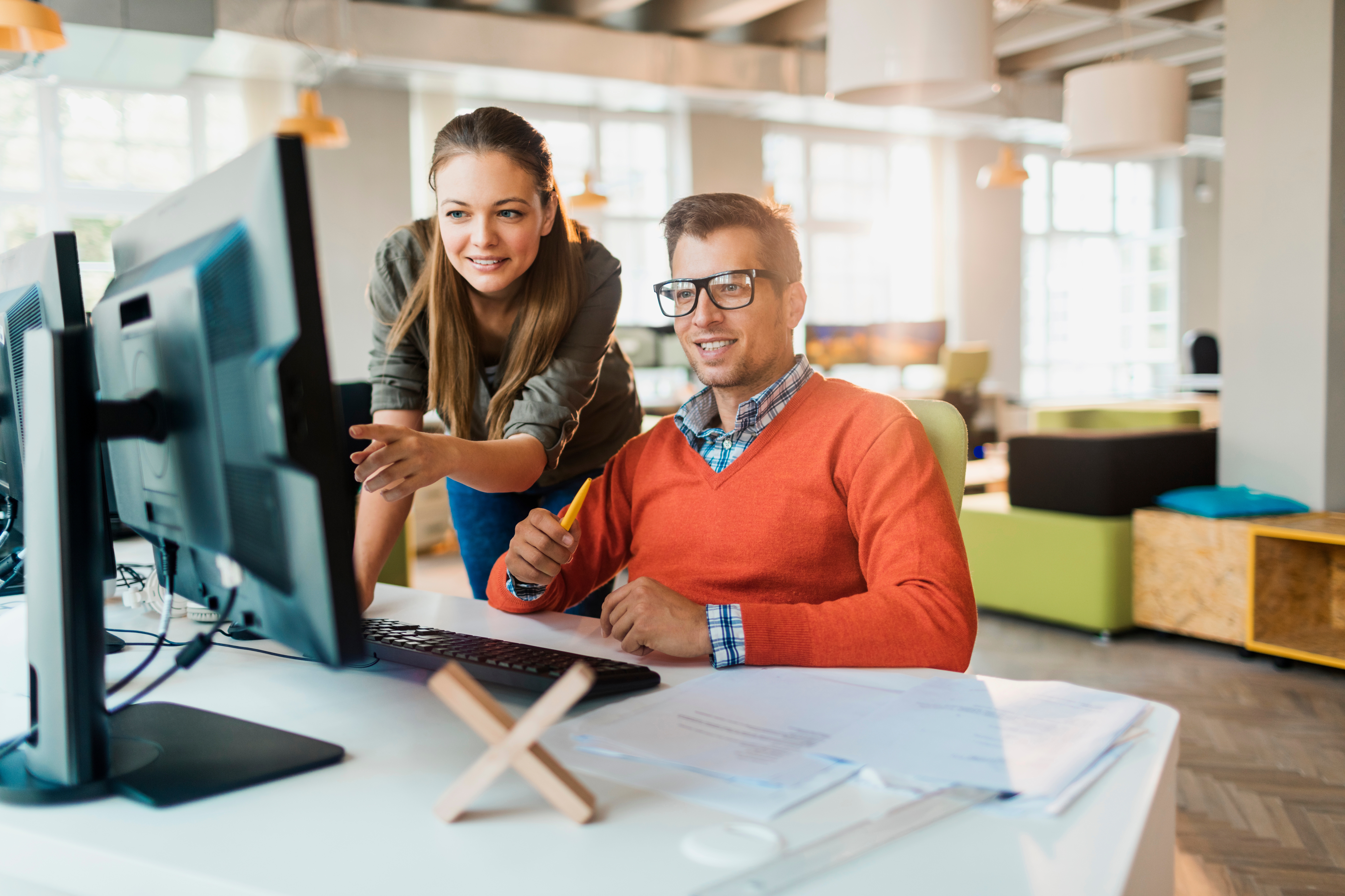 Two colleagues work together in a modern office. They are reviewing a website on a large monitor while pointing to particular features on the screen.