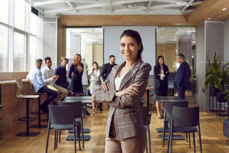 A smiling woman stands with her arms crossed in a corporate training room. She’s confident because she attended our professional writing courses and learned to communicate effectively.