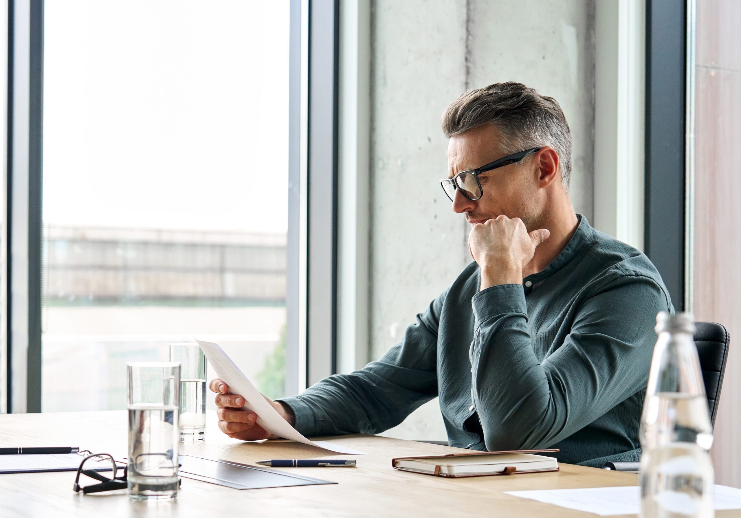 A man sits in an office reading board papers with his hand on his chin - he is concentrating and looking thoughtful