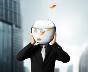 A person in a business suit has a goldfish bowl instead of a head. A goldfish is jumping out of the bowl.