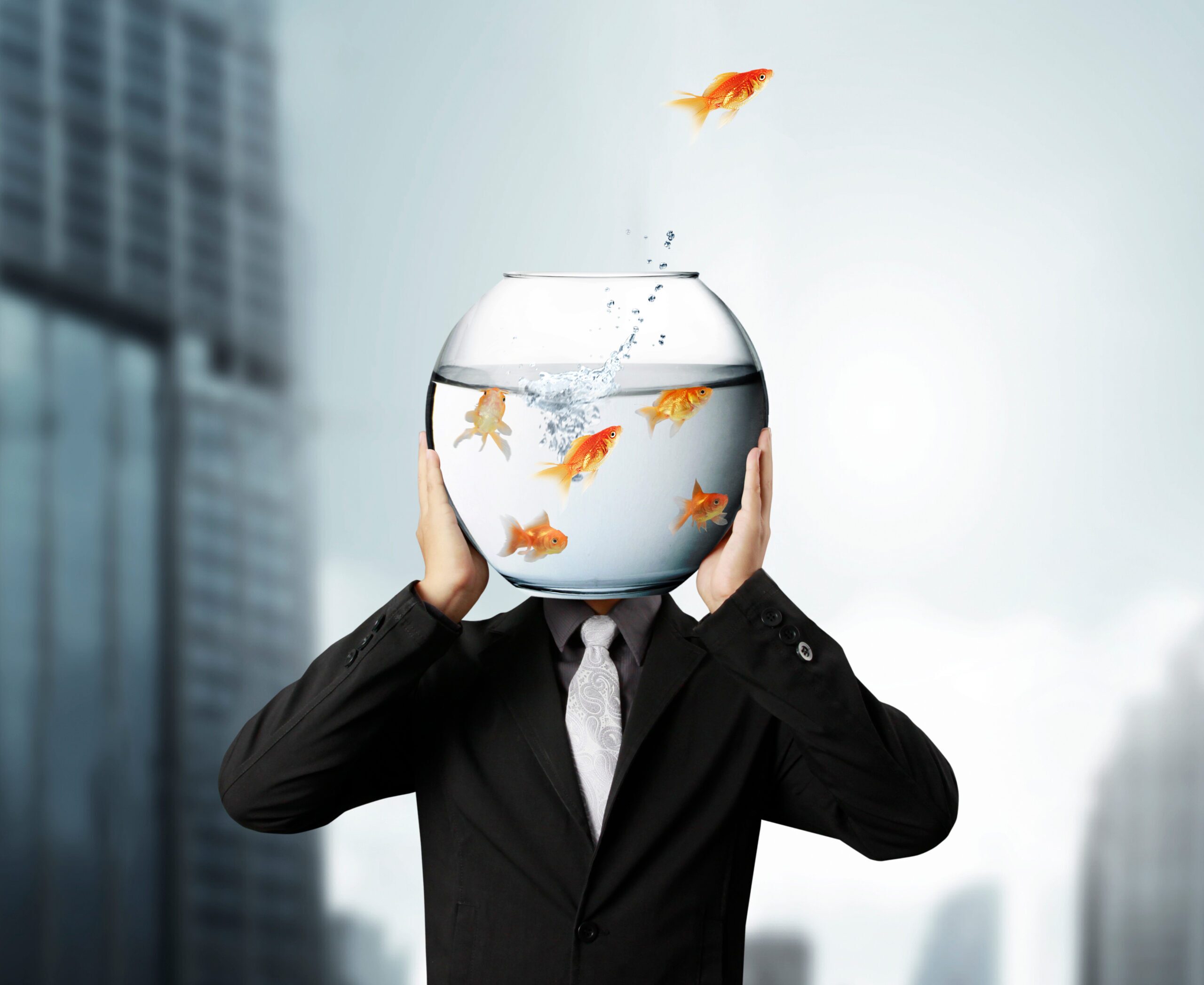 A person in a business suit has a goldfish bowl instead of a head. A goldfish is jumping out of the bowl.