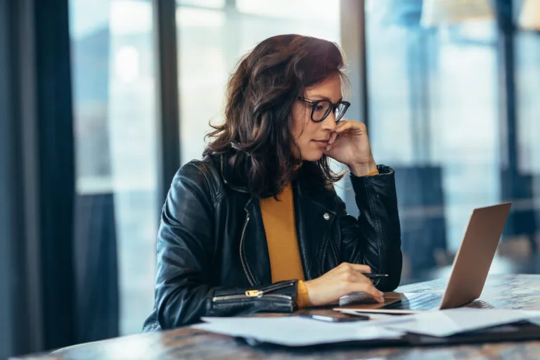 A woman in a leather jacket works on her laptop. She’s writing to inform her readers.