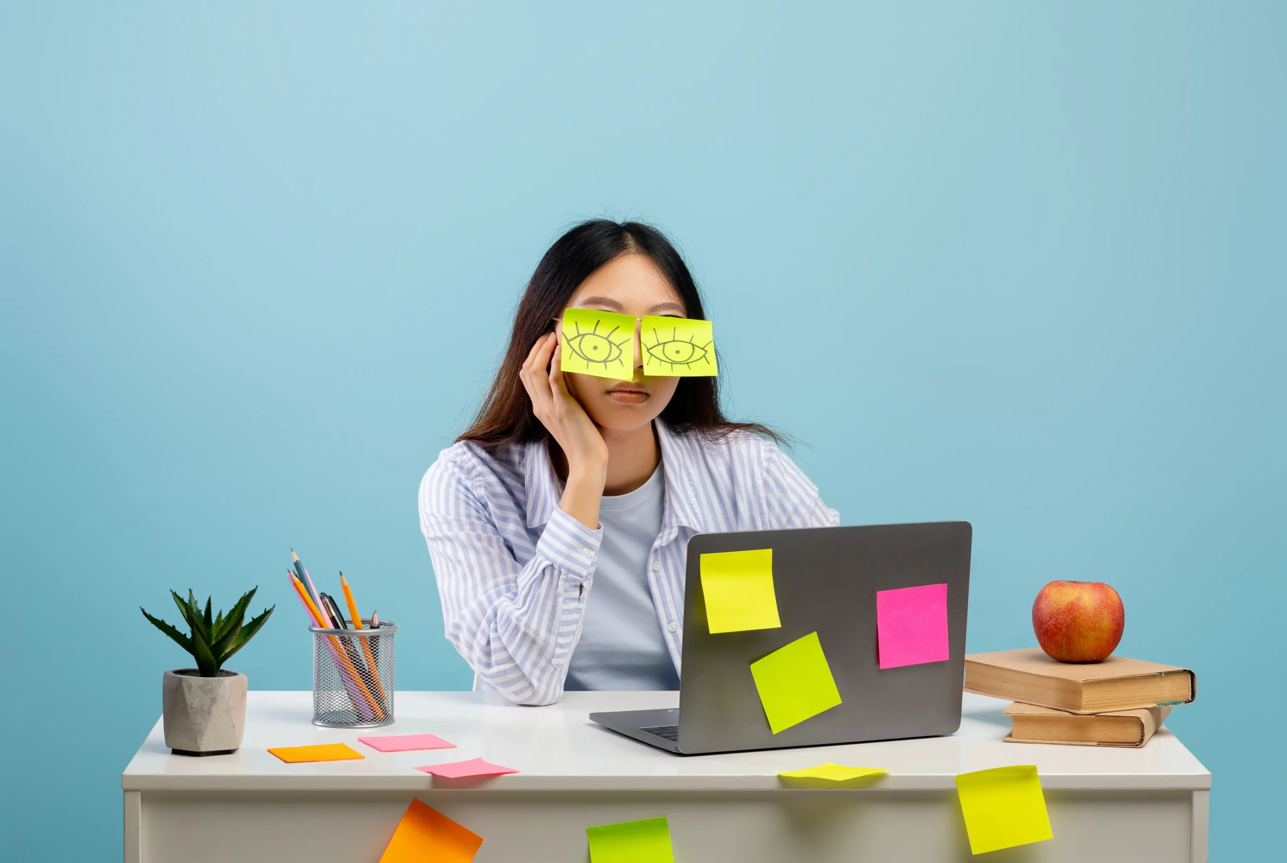 Office worker sits despondently at her laptop. She’s stuck two sticky notes with eyes drawn on them over her eyes.